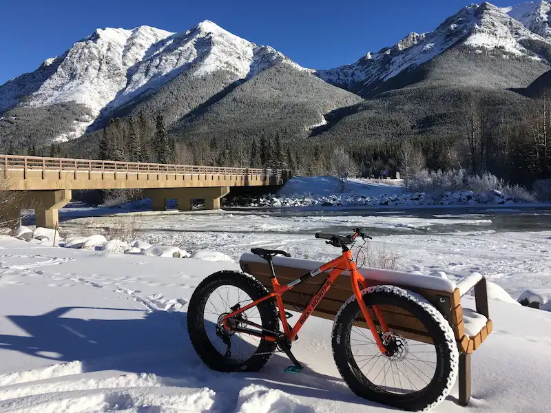 Image of a fat mountain bike resting against a bench in the winter at the Canadian Rockies with mountains and a bridge at the background.