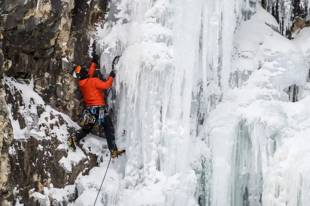 Image of man ice climbing in Waterton National Park in winter clothes with climbing gear on him such as a mountaineering axe, carabiners, and rope to climb frozen icycles up a cliff face.