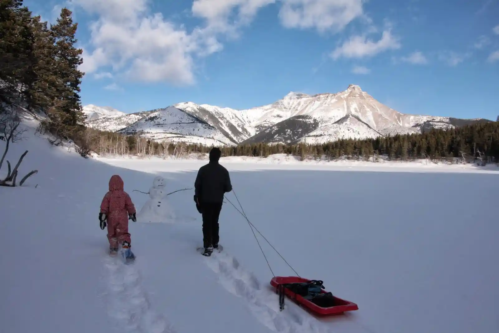 Image of a man and his daughter wearing winter clothing walking through a snowfield in Waterton National Park while bringing a sled with beautiful, scenic, and dramatic mountains in the background.