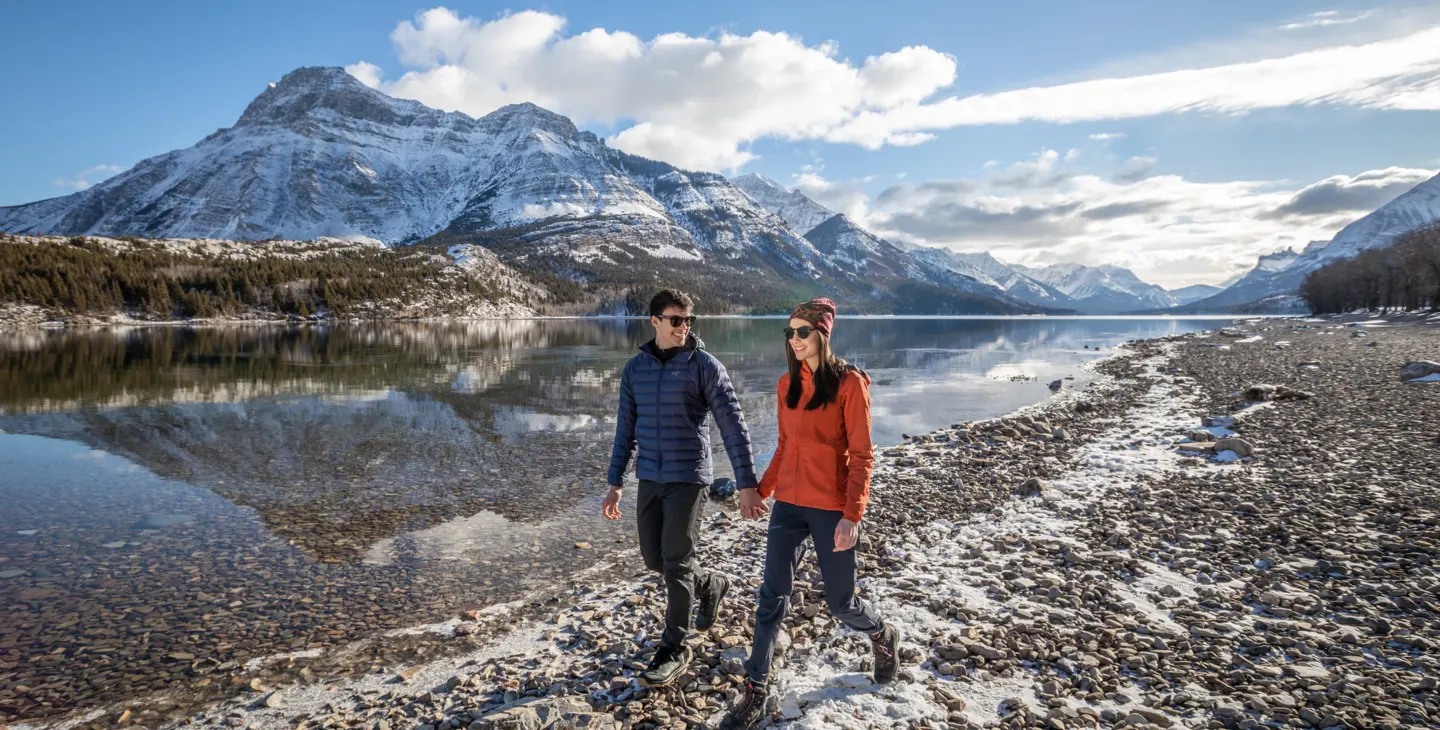 Image of man and woman woman alongside the main lake of Waterton National Park with the lake and mountains at the background